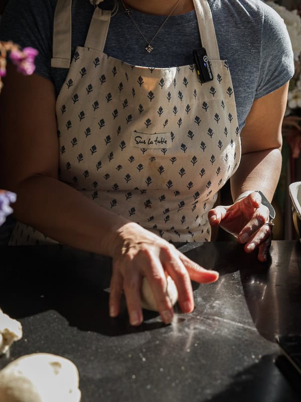 A person wearing a patterned apron is kneading dough on a dark countertop in a sunlit kitchen. Their hands are positioned over the dough, and they are wearing a smartwatch on their left wrist.